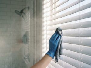 Gehandschoende hand reinigt witte horizontale jaloezieën met microvezeldoek in moderne badkamer met waterdruppels