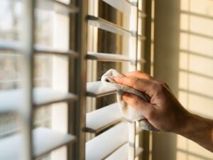 Hand veegt witte horizontale jaloezie schoon met microvezeldoek, stofdeeltjes zichtbaar in zonlicht