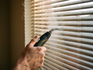 Hand houdt stoomreiniger vast die damp op jaloezieën spuit met waterdruppels en zonlicht door raam