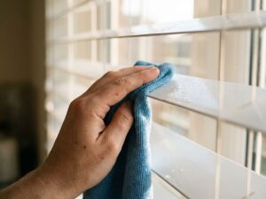 Hand reinigt verticale witte jaloezie met microvezeldoek, waterdruppels zichtbaar op lamellen in natuurlijk daglicht