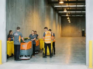 Professioneel schoonmaakteam in werkkleding reinigt grote lege bedrijfsruimte met industriële schrobmachines op betonnen vloer.