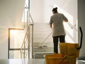 Professionele schoonmaker dweilt marmeren trappenhuis van appartementencomplex met gele emmer en mop op de voorgrond.