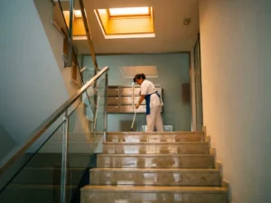 Professionele schoonmaker dweilt een strakke trappenhaal van een appartementencomplex, met glanzende tegels en metalen brievenbussen op de achtergrond.