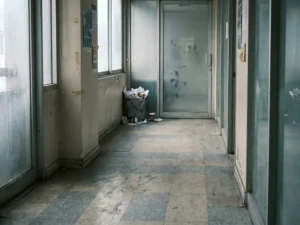 Verwaarloosd kantoorgangpad met vuile tegelvloer, overloopende prullenbak en besmeerde glazen deur in gedempte grijstinten.